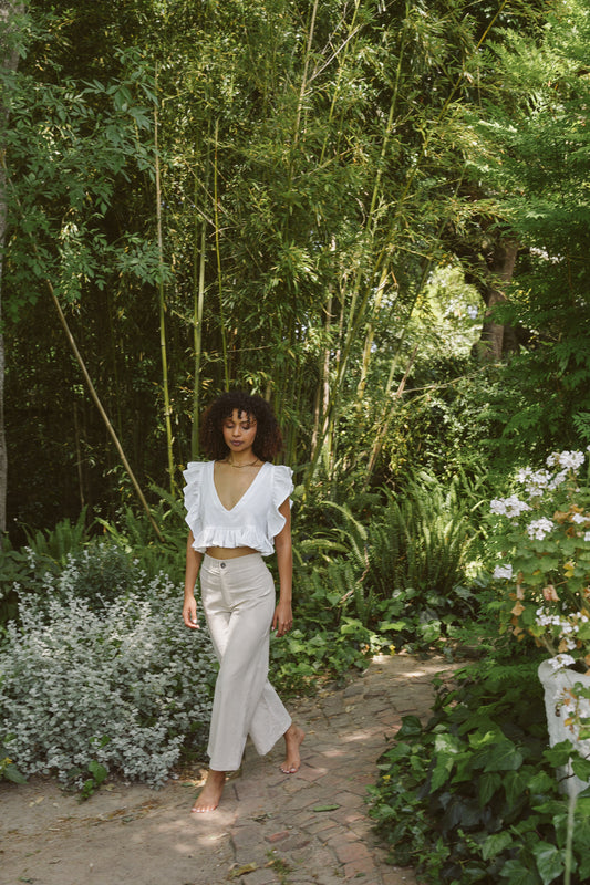Woman in a white frilly top and oatmeal linen cotton pants standing in a garden.