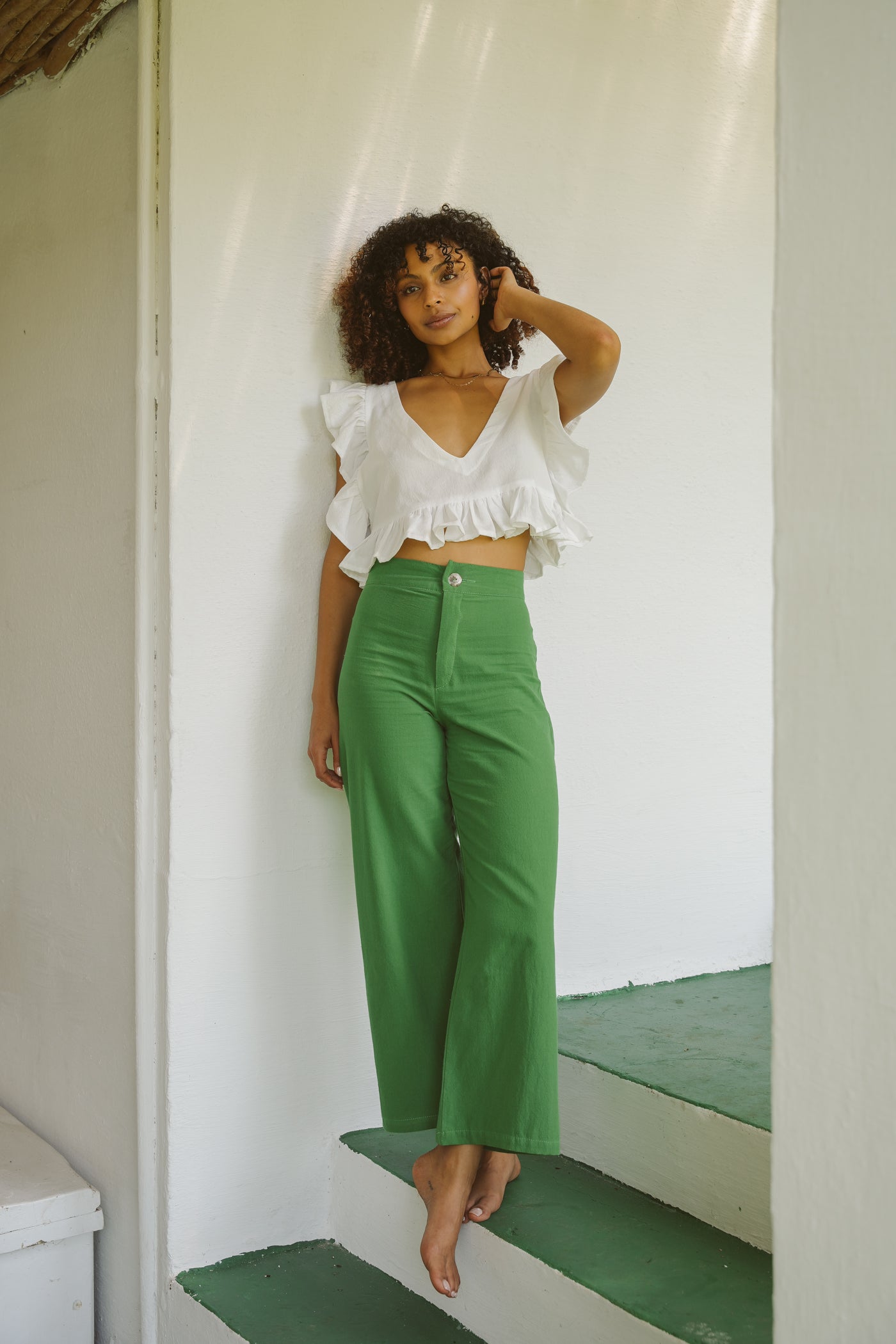 Woman in a white frilly cotton top and jade green coloured cotton trousers standing on a green staircase.