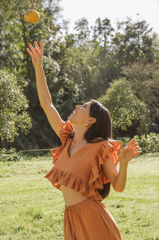 Woman in our clay frilly top and drawstring pants reaching up to catch an apple in a park.
