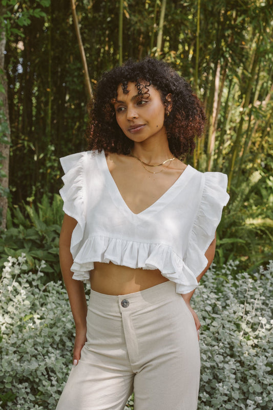 Woman wearing a white Frilly Top and oatmeal Barley Trousers standing in a garden.