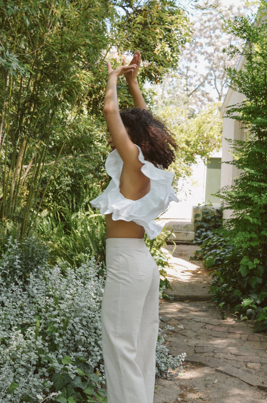 Woman in a white Frilly Top and Oatmeal Barley Trousers standing in a garden with greenery and flowers.