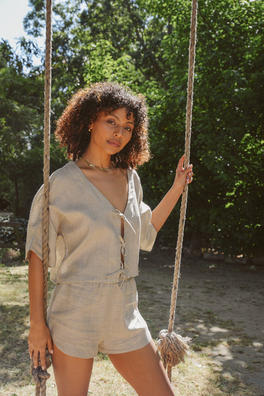 Woman in a oatmeal linen native top and beth shorts sitting on a swing with greenery in the background