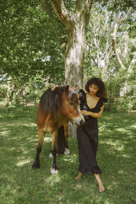 Woman in a long black Poppy dress standing next to a horse in a park-like setting with trees and grass.
