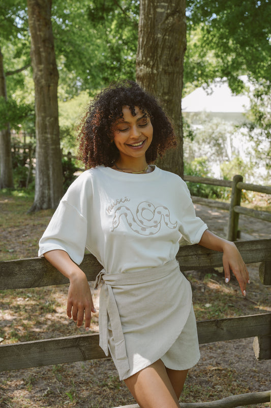 Woman wearing a white Serpent embroidered t-shirt with embroidery and beige Cherie cotton skirt standing outdoors near trees.
