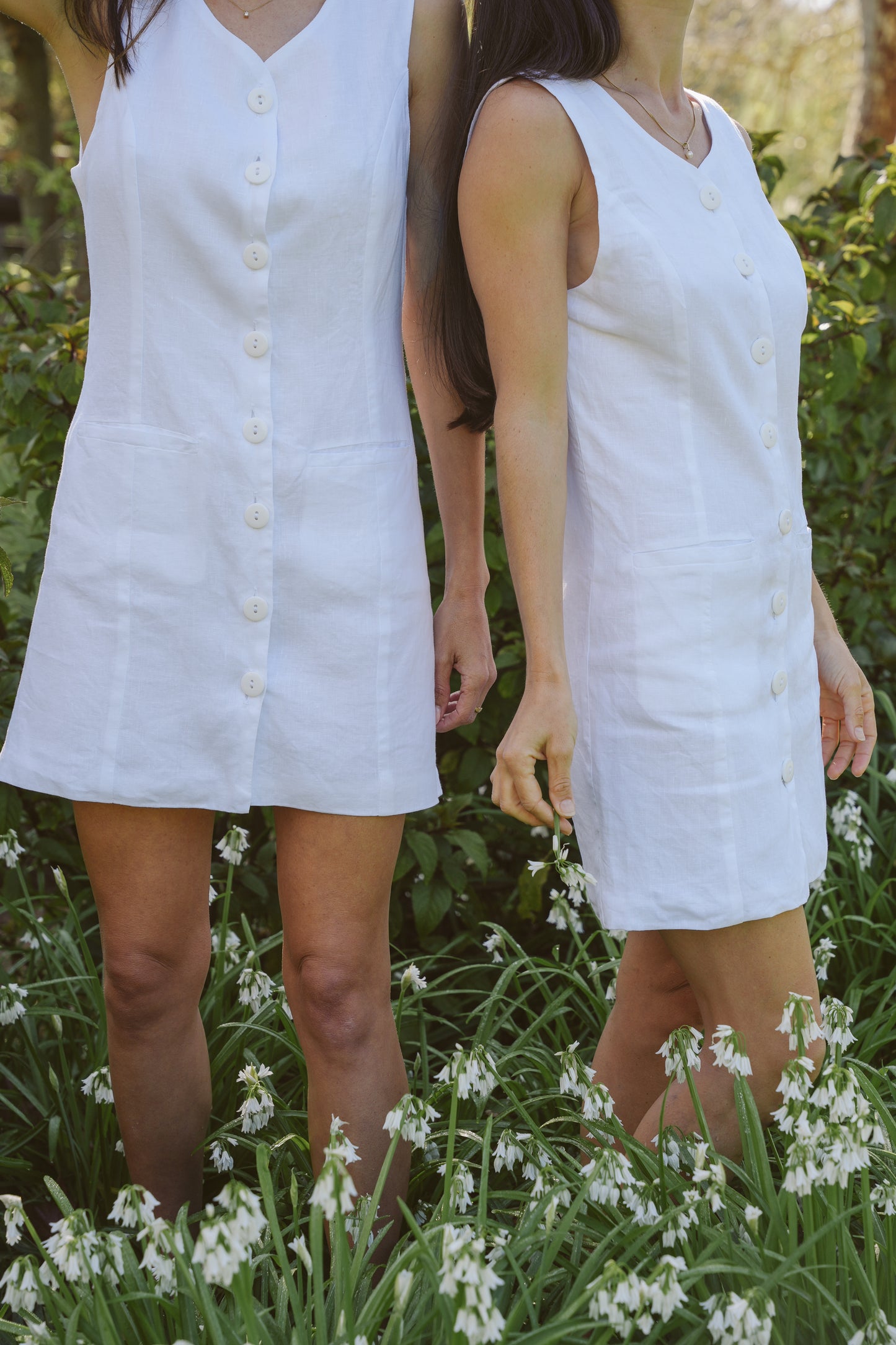 Two women wearing white linen sleeveless dresses standing in a garden with greenery and flowers.