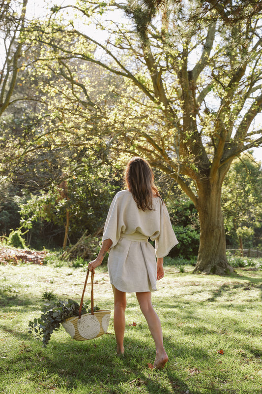 Woman in a oatmeal linen wrap dress walking through a garden with trees and greenery.