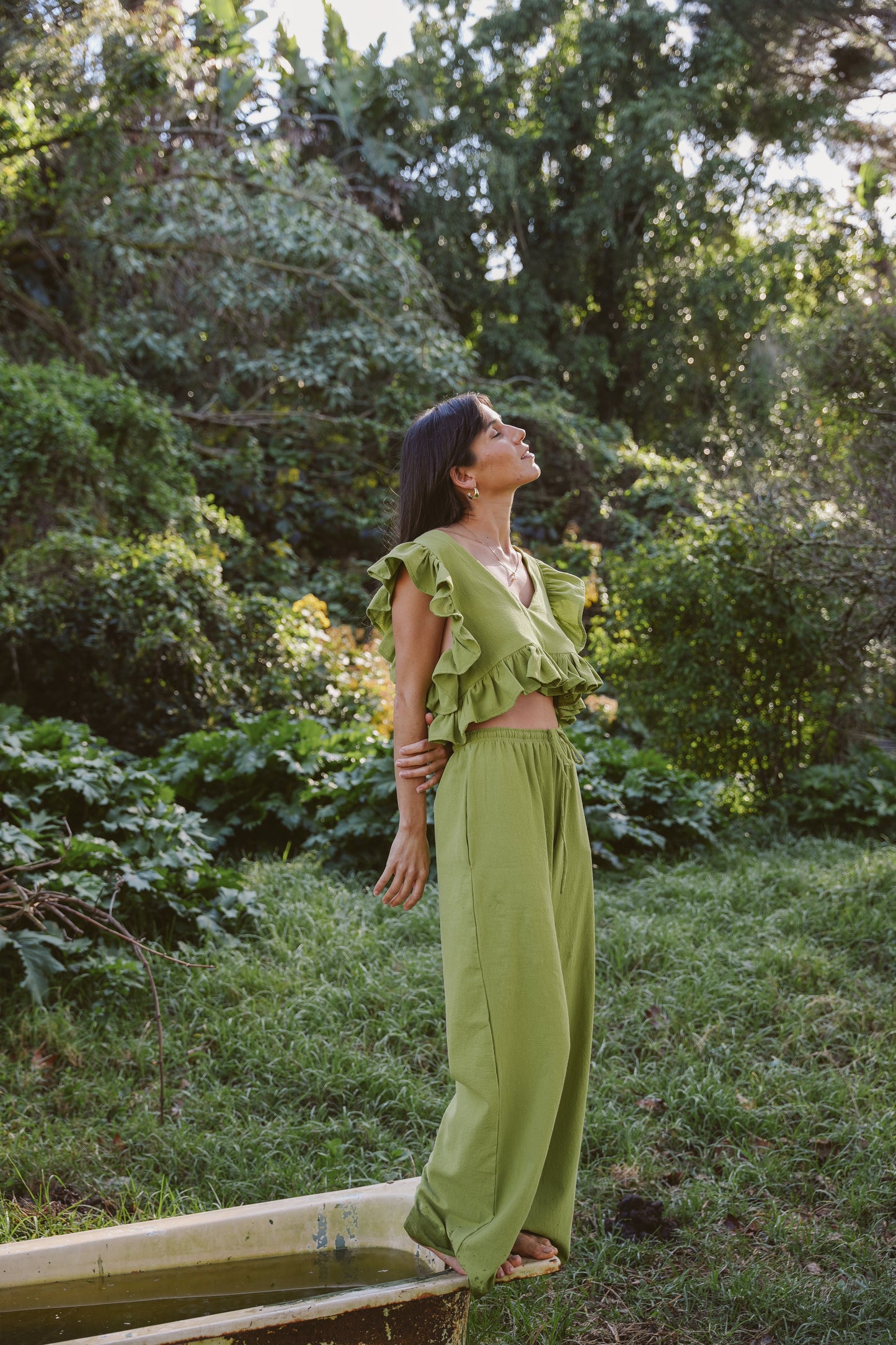Woman in a wasabi green frilly top and drawstring cotton pants standing in a garden