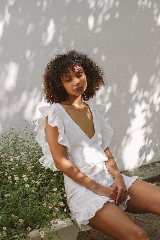 Woman in a white cotton outfit sitting outdoors against a light-colored wall.