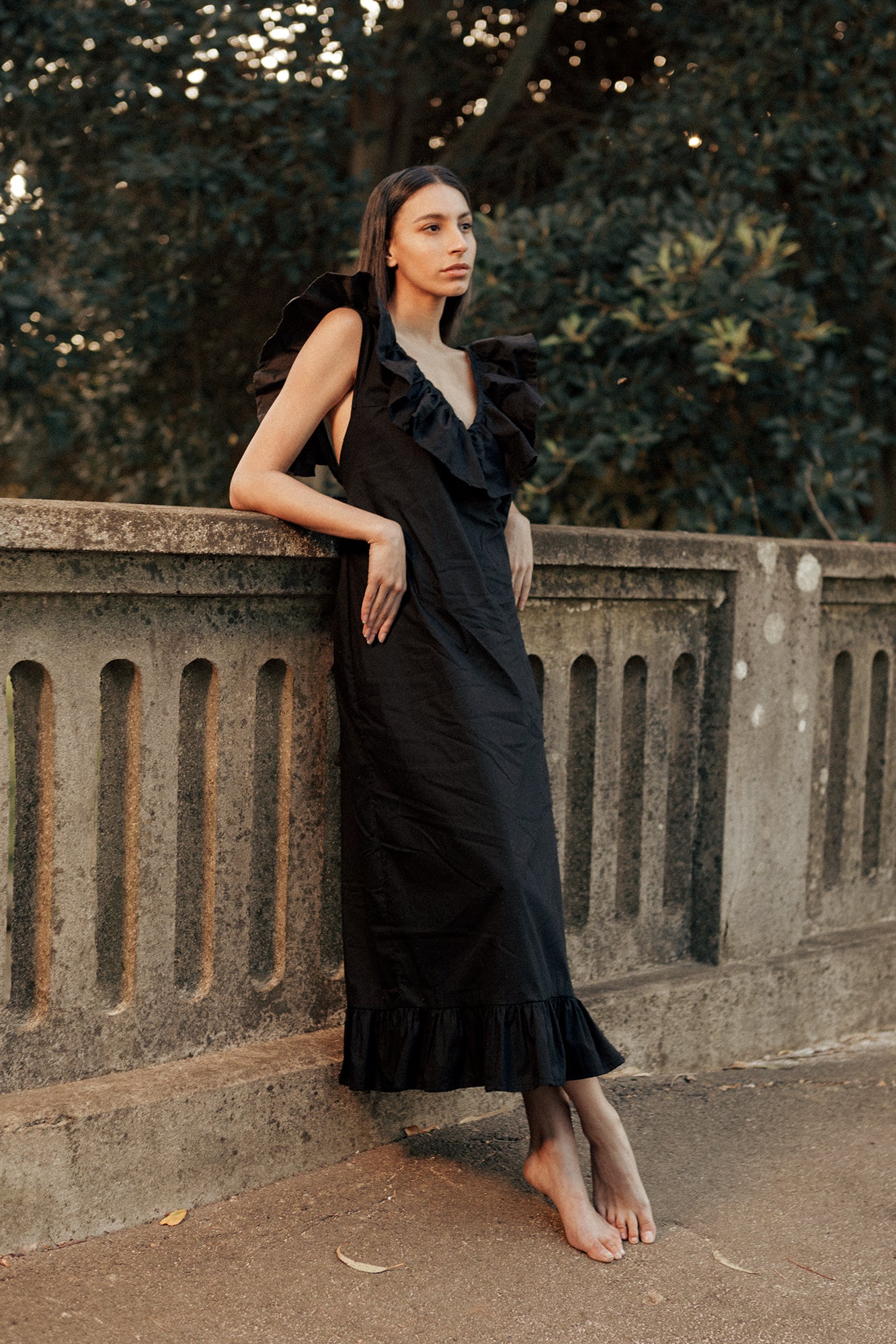 Model wearing a black Museā Poppy Dress that is maxi length. Model is posing on a bridge in the forest.