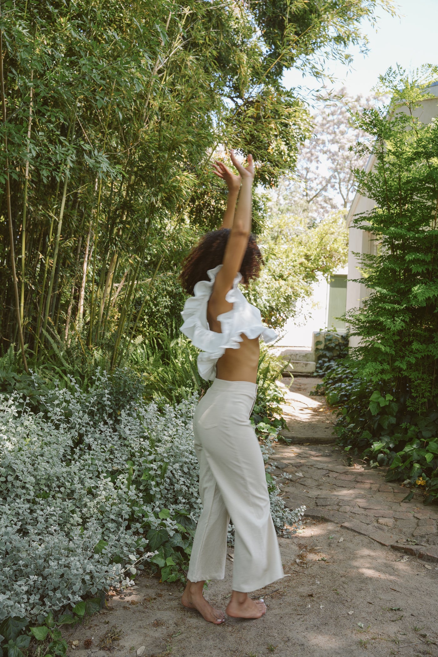 Woman in a white frilly top and oatmeal trousers standing in a garden with greenery and a path.