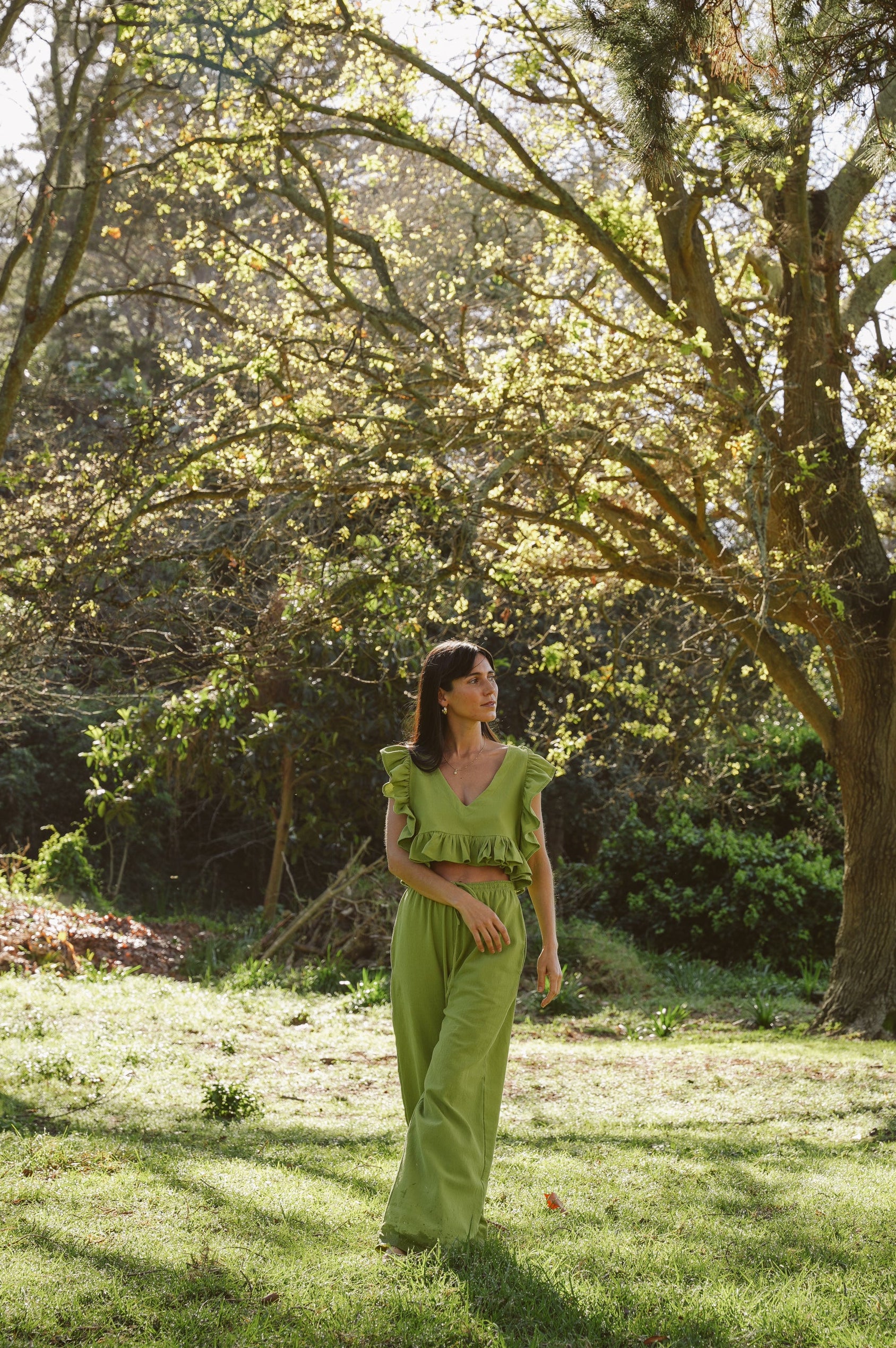 Woman in a wasabi Frilly Top and drawstring trousers standing in a sunlit garden with trees and grass.