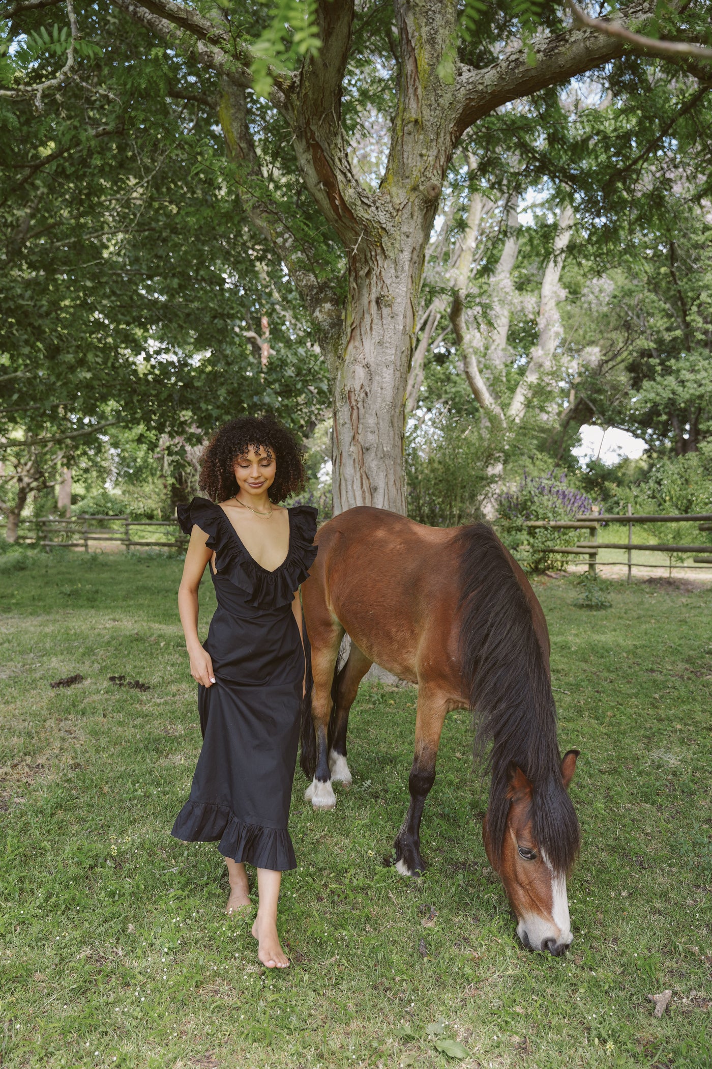 Woman in a black Poppy dress standing next to a horse in a grassy field with trees in the background
