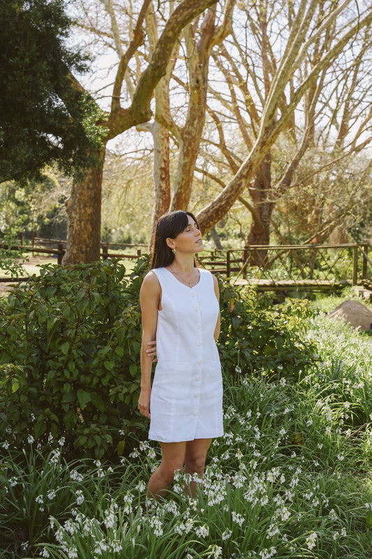 Woman in a white linen shift dress standing in a garden with trees and flowers.
