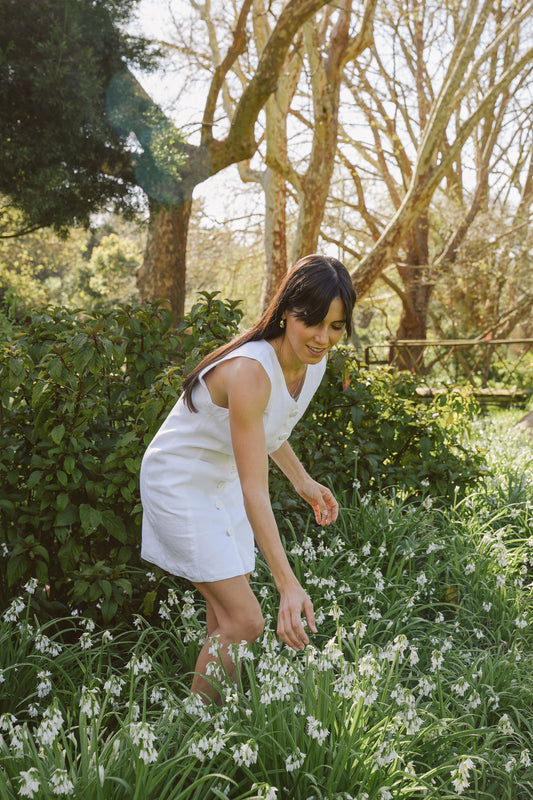 Woman in a white linen shift dress standing in a garden with greenery and flowers.