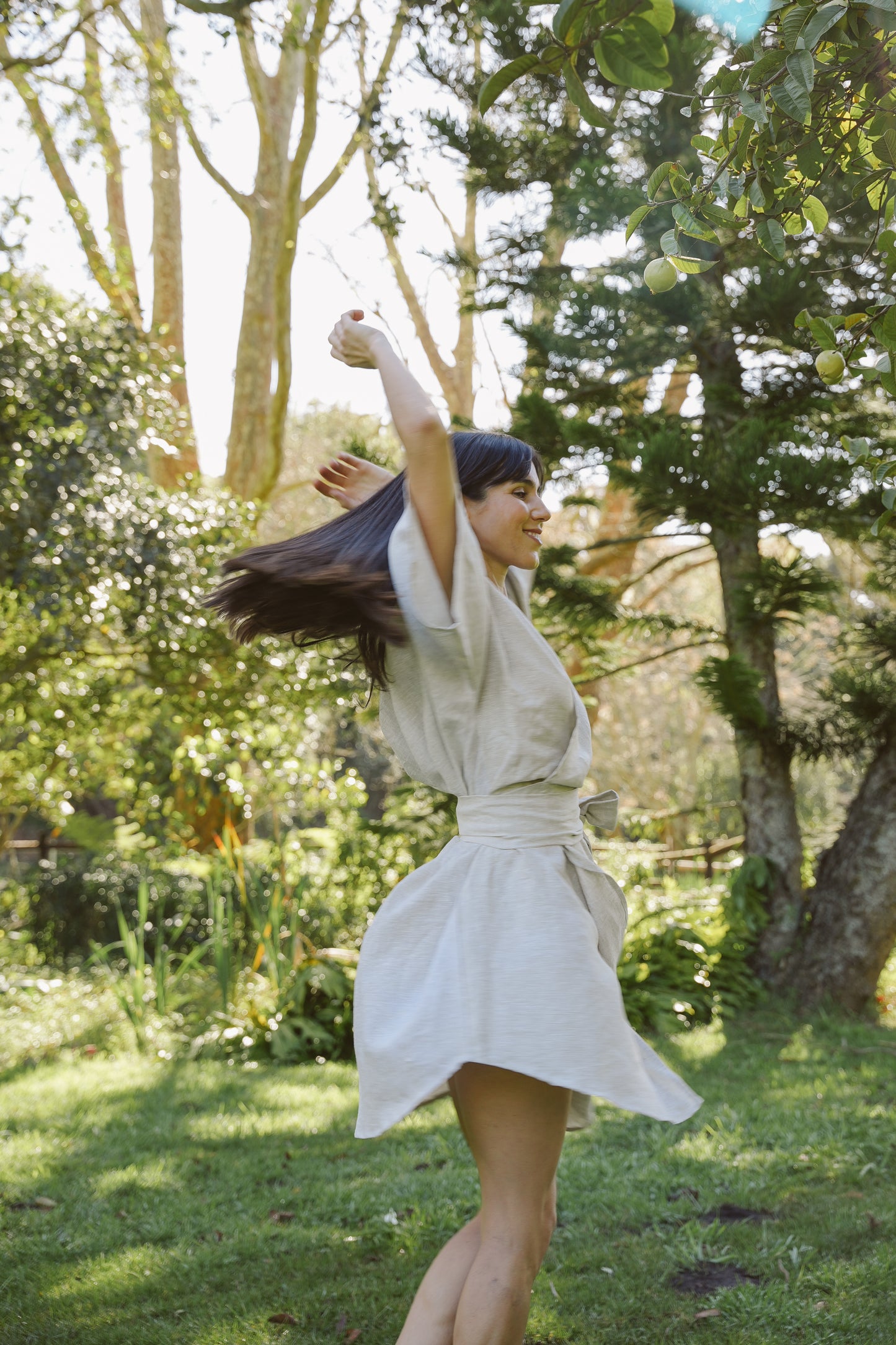 Woman in a oatmeal wrap linen dress dancing in a park with trees and grass in the background
