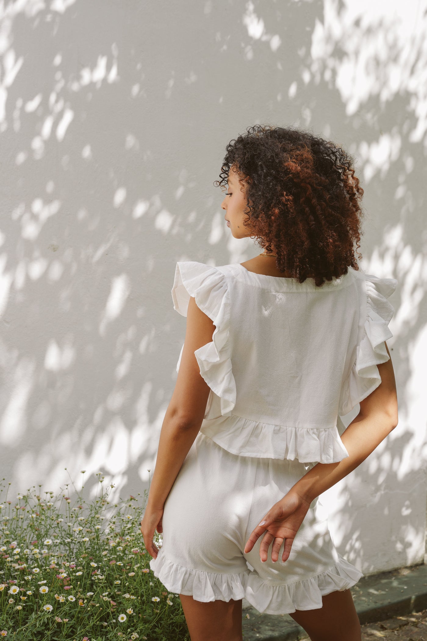 Woman wearing a white frilly top and prancing cotton shorts against a white wall outdoors